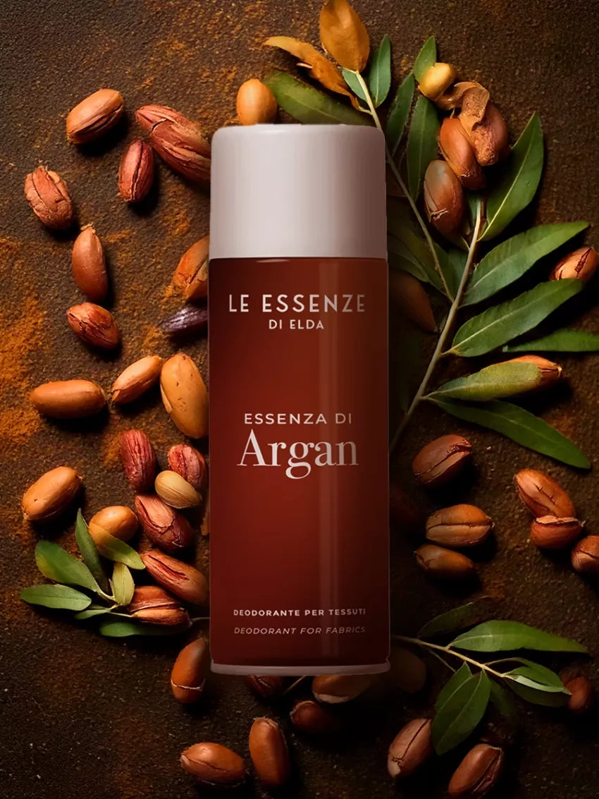 Argan textílsprey 400ml frá Le Essenze di Elda í rauðbrúnum úðabrúsa með hvítu loki, umkringt hnetum og grænum laufblöðum á brúnum bakgrunni.